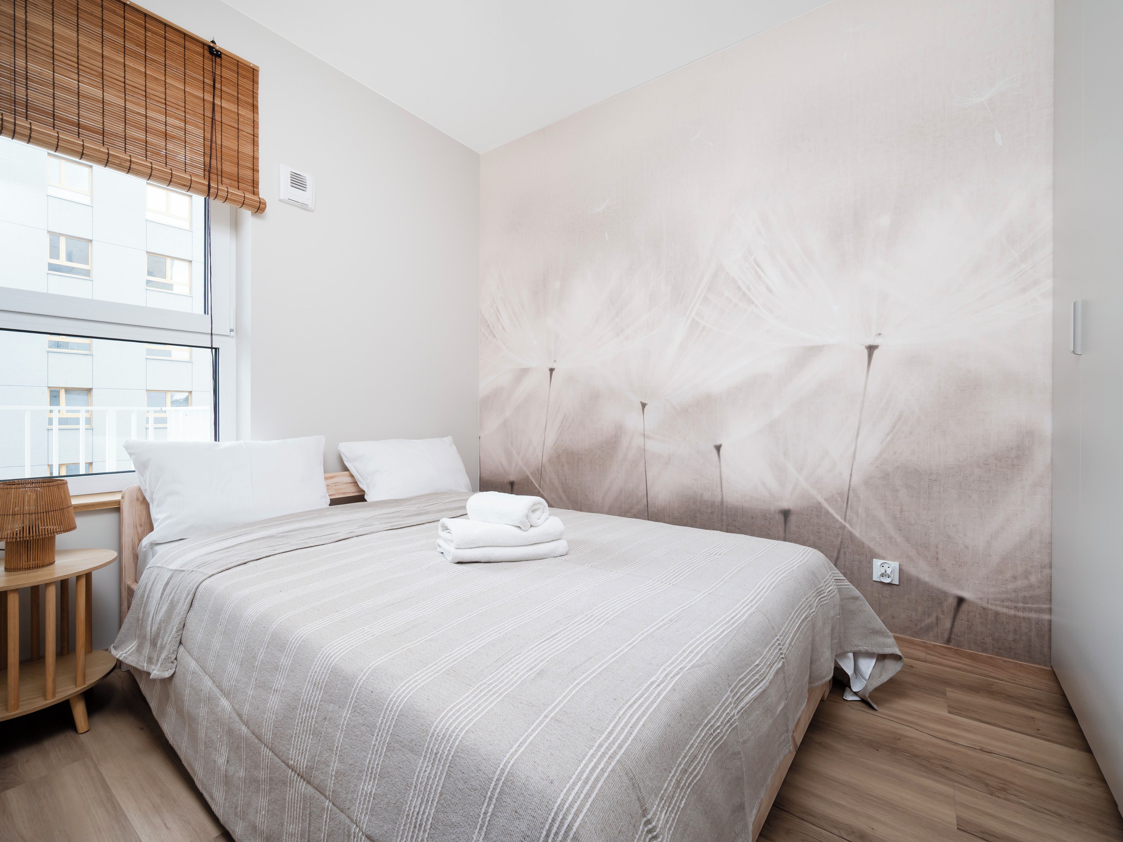 Ein Hotelzimmer mit einem großen Bett, einem Holznachttisch, einem Fenster mit Jalousien und einer Wand mit strukturierter Oberfläche.