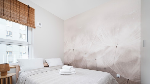 Ein Hotelzimmer mit einem großen Bett, einem Holznachttisch, einem Fenster mit Jalousien und einer Wand mit strukturierter Oberfläche.