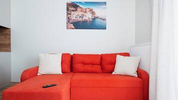 Um sofá seccional vermelho com um controle remoto, uma almofada branca e listrada, e uma imagem de uma cidade costeira na parede.
