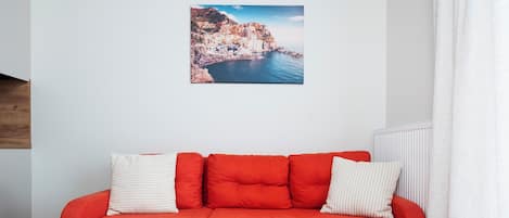 A red sectional sofa with a remote control on it, a white and striped pillow, and a picture of a coastal town on the wall.