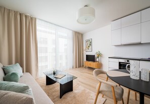 Una sala de estar moderna con una mesa de centro de vidrio, un sofá con cojines, una zona de comedor con una silla, una cocina con armarios y un ventanal con cortinas.