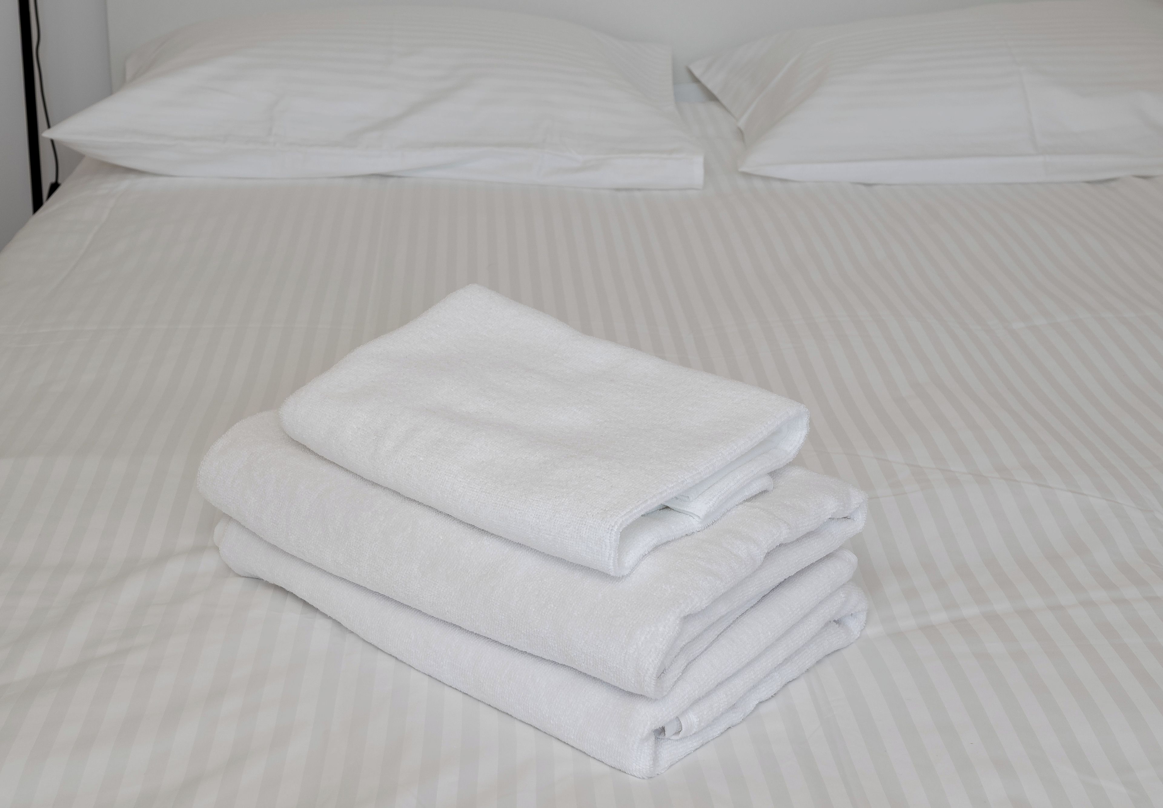 A neatly made bed with white bedding and pillows, and a stack of folded white towels on the bed.