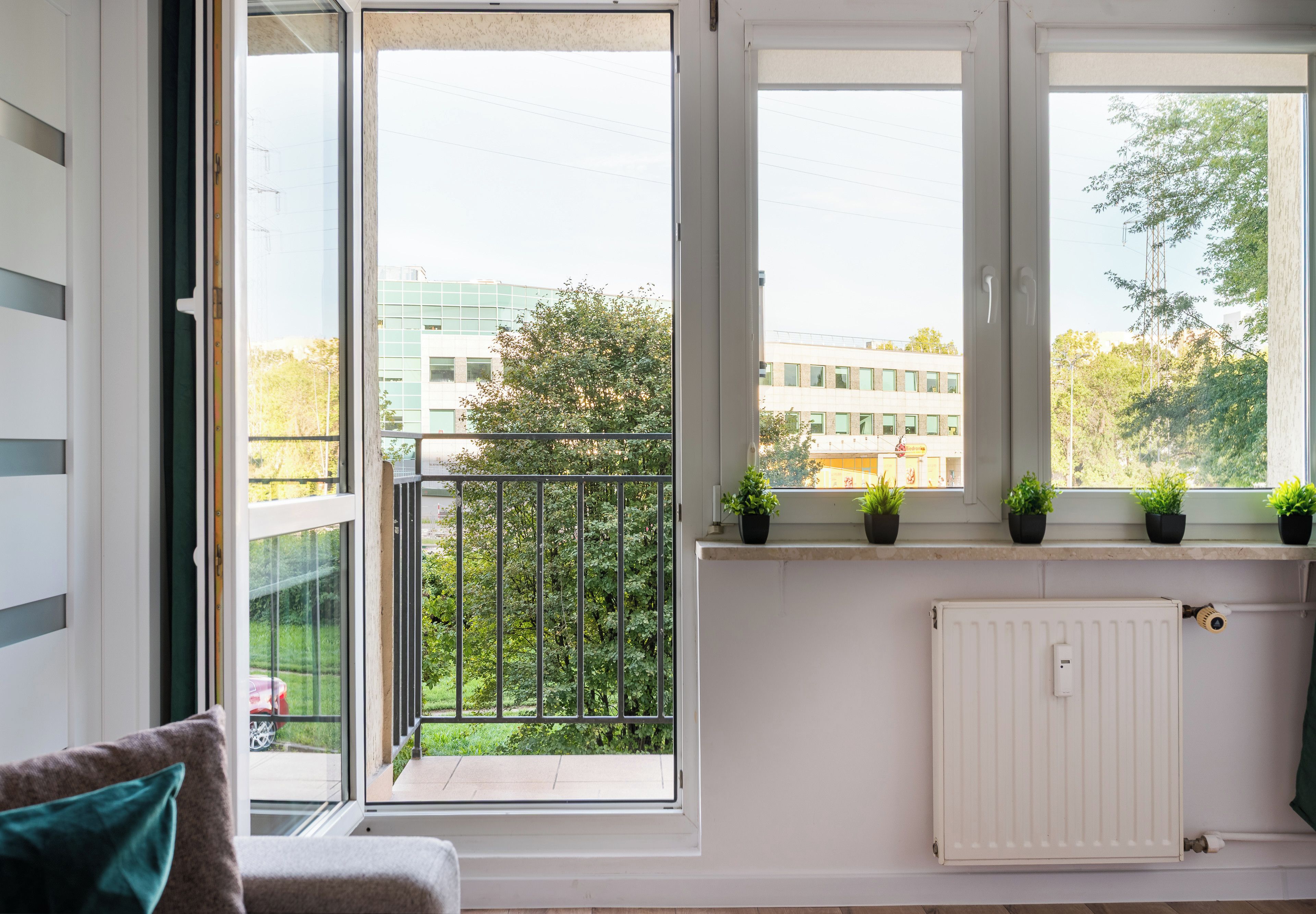 Ein Zimmer mit Blick auf ein Gebäude und Bäume durch große Fenster, inklusive Heizkörper und einer Couch mit Kissen.
