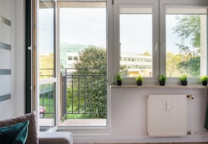 A room with a view of a building and trees through large windows, a radiator, and a sofa with a cushion. - Mokotów Rzymowskiego Urban Apartment (Warszawa)
