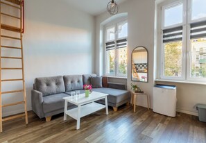 A modern living room with a grey sofa, a white coffee table, a ladder, and a window with a view of buildings. - Piastów Loft Mezzanine Studio (Szczecin)