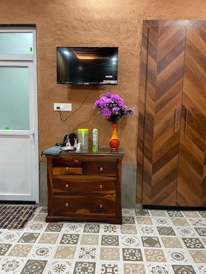 A room with a wooden cabinet, a television mounted on the wall, a vase with purple flowers on a glass table, and patterned floor tiles.
