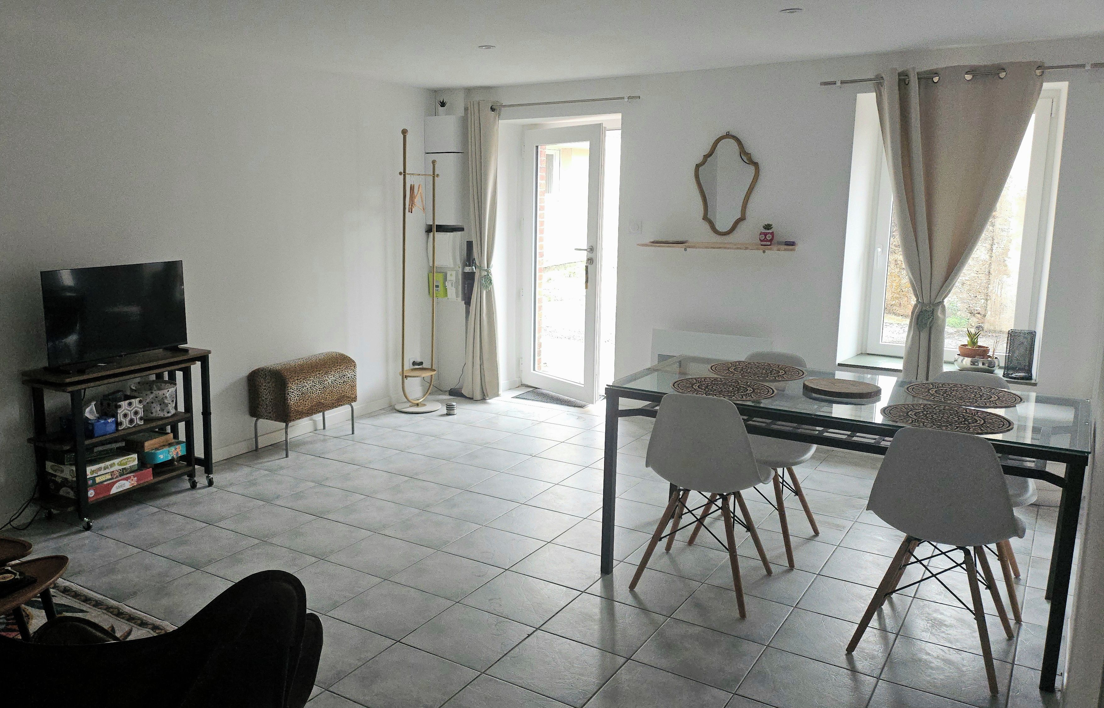 Een woonkamer met een televisie, een eettafel met stoelen, een bank en een raam met gordijnen.