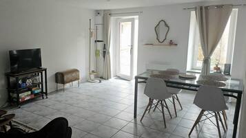 Een woonkamer met een televisie, een eettafel met stoelen, een bank en een raam met gordijnen.
