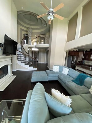 A spacious living room with a sectional sofa, a fireplace, a television, and a staircase leading to an upper level. - Great Family Escape | Pool, Hot Tub + Movie Room (Pearland)