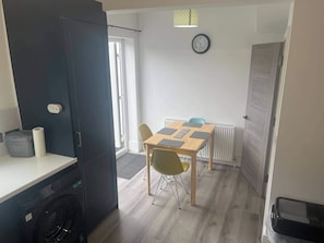 A compact kitchen with a washing machine, a dining area with a table and chairs, and a clock on the wall. - The 139 Sleeps 5 - Garden - Free Parking (St. Leonards-on-Sea)
