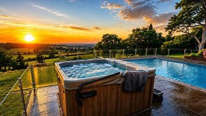 Ett bubbelbad med träpanel runt om, omgivet av en swimmingpool, med utsikt över en solnedgång och ett vackert landskap.