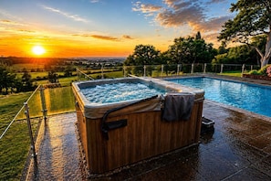 A hot tub with a wooden exterior, surrounded by a swimming pool, with a view of a sunset and landscape. - Luxury Cottage Escape Sleeps 4 with Hot Tub & Pool (Keston)