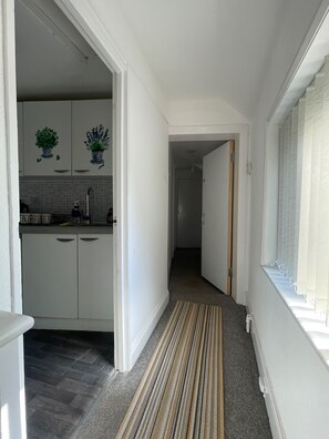 En smal hall med ett köksområde på vänster sida, med skåp och en kakelbeklädd vägg bakom arbetsbänken. En randig matta löper längs hallens mitt och leder fram till en dörr i slutet.