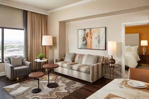 A hotel room with a sofa, armchair, dining table, and a large window with curtains.