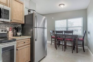 A modern kitchen with a stainless steel refrigerator, a microwave, and a coffee maker. There is a dining table with chairs and a window with blinds. - The Grounded Oak | 1mi to UT, Fenced Yard (Tampa)