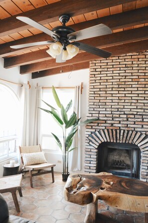 A cozy living room with a brick fireplace, a ceiling fan, and a large plant. - Playa Plateada Beach House (Ensenada)