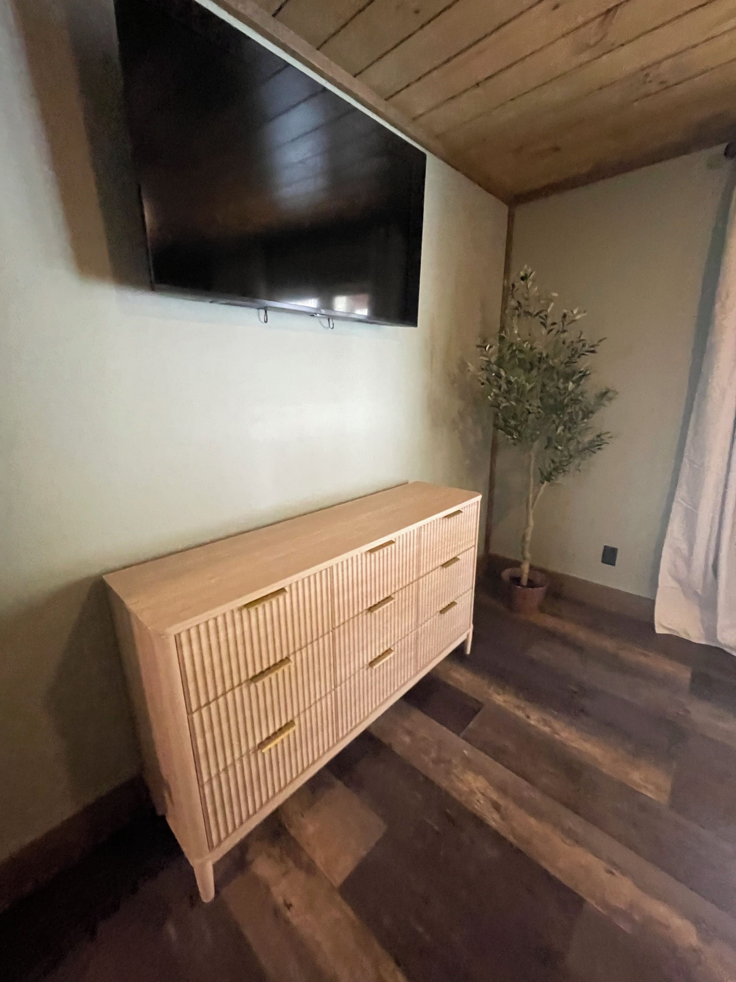 Een kamer met een houten vloer, een lichtkleurige kast, een potplant en een flatscreen televisie aan de muur.