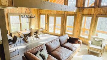 Een ruime binnenruimte met een hoge plafond, houten balken, een grote bruine leren bank, een eettafel met stoelen en grote ramen met uitzicht op het omliggende landschap.