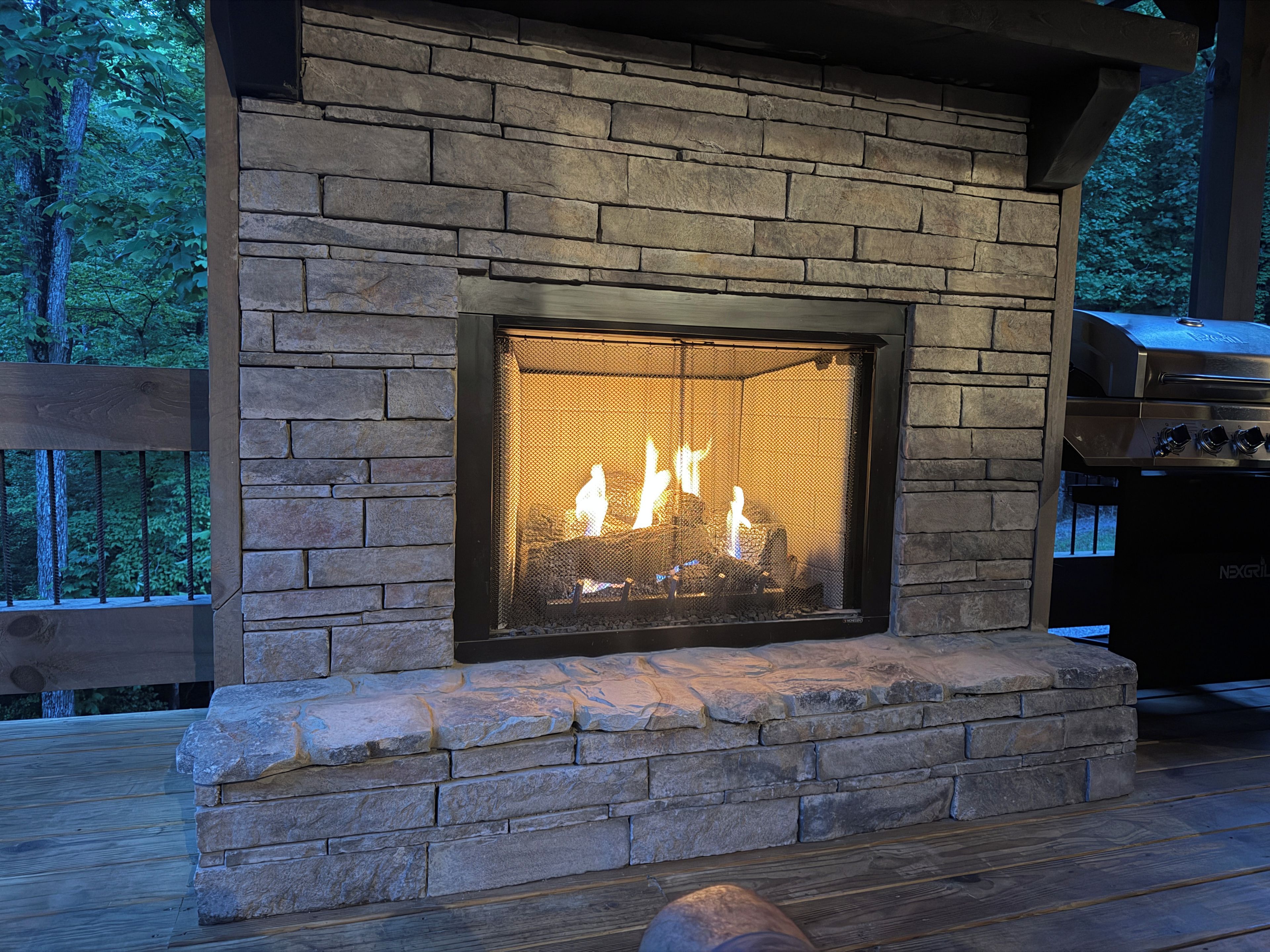 A stone fireplace with a glass screen, set in a wooden deck with a view of trees.