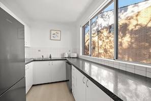 A kitchen with white cabinets, a black countertop, and a window overlooking a brick building. - Stylish 3 bedroom apartment in Miranda steps to Westfield and train station. (Miranda)