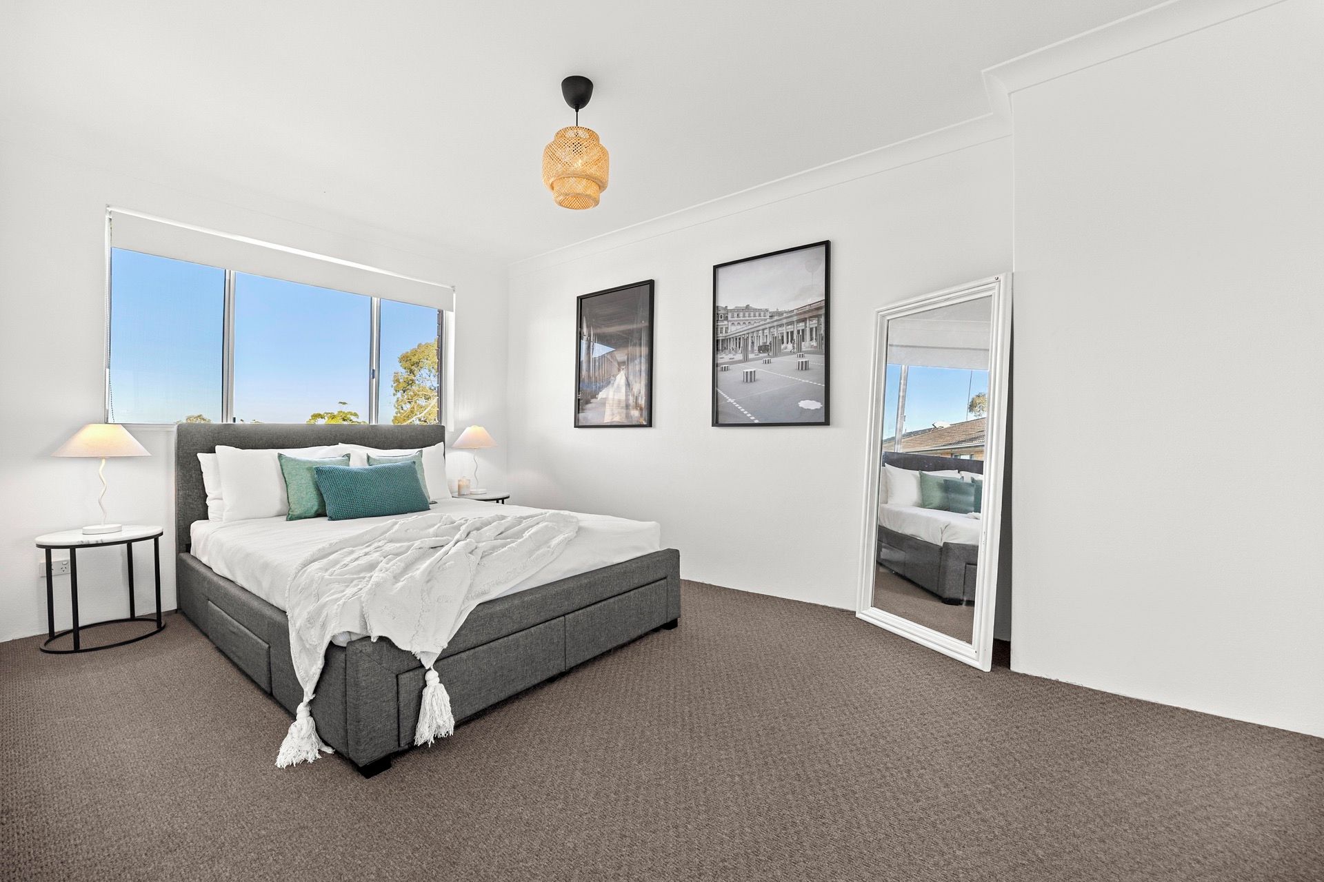 A modern bedroom with a large bed, two bedside tables, a hanging light fixture, and a view through a door.