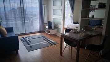 A modern living room with a sofa, a dining table, and a kitchen area with a bed and a window.