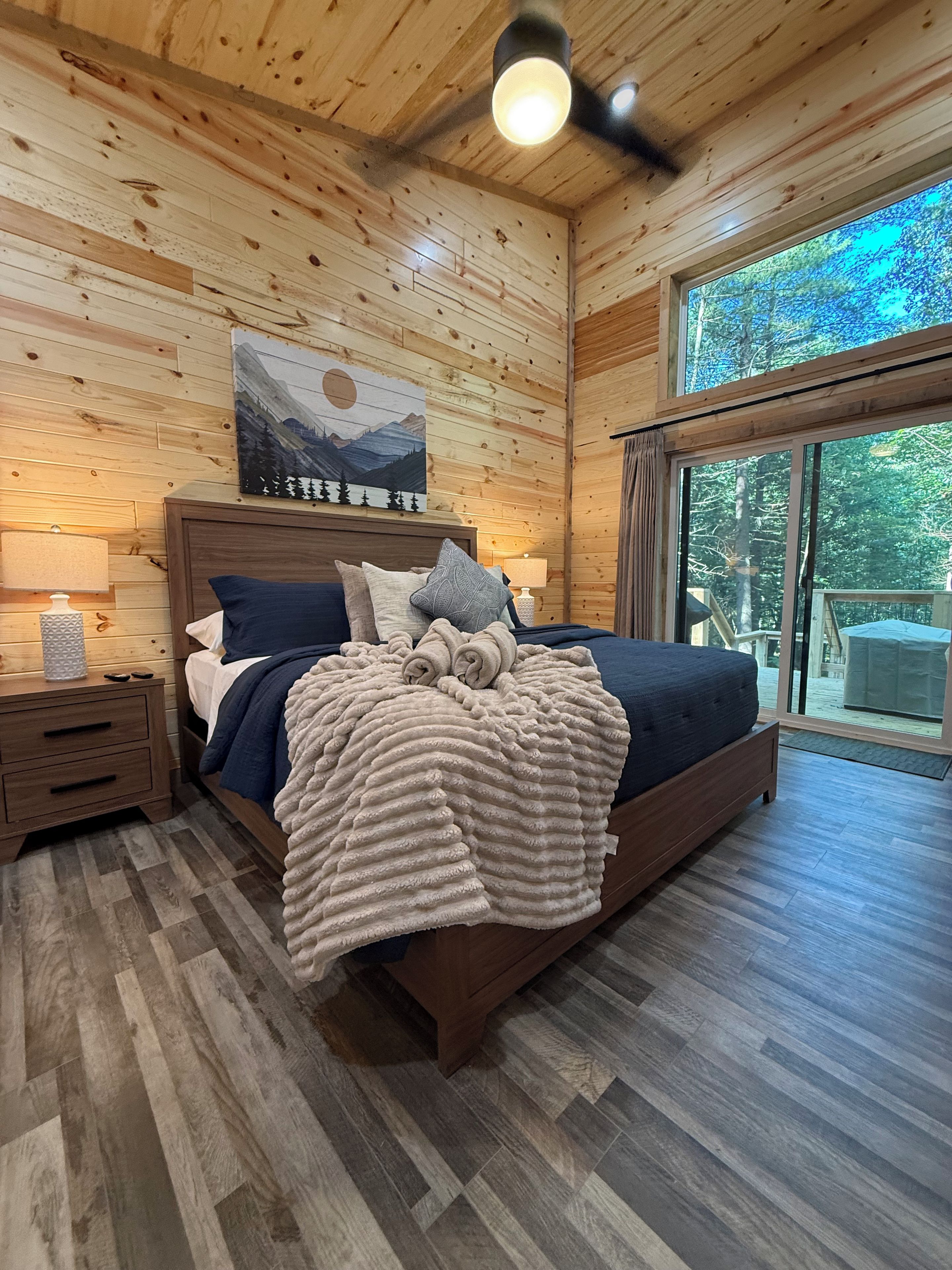A wooden-paneled bedroom with a bed, a nightstand, a lamp, and a painting on the wall.