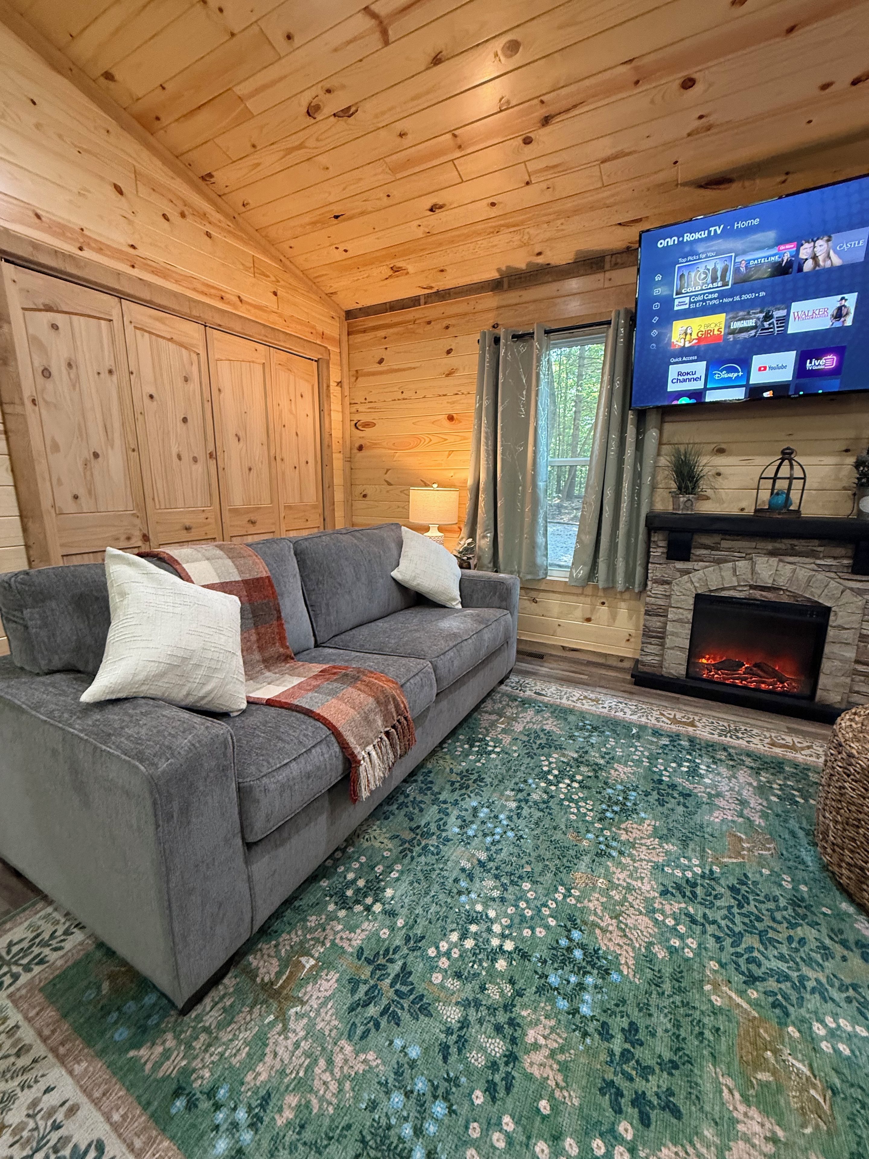 A cozy cabin interior with a gray sofa, a patterned rug, a fireplace, and a television displaying a Roku interface.