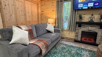 A cozy cabin interior with a gray sofa, a patterned rug, a fireplace, and a television displaying a Roku interface.
