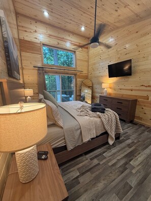 A cozy bedroom with a bed, a TV, a window, and a wooden floor.