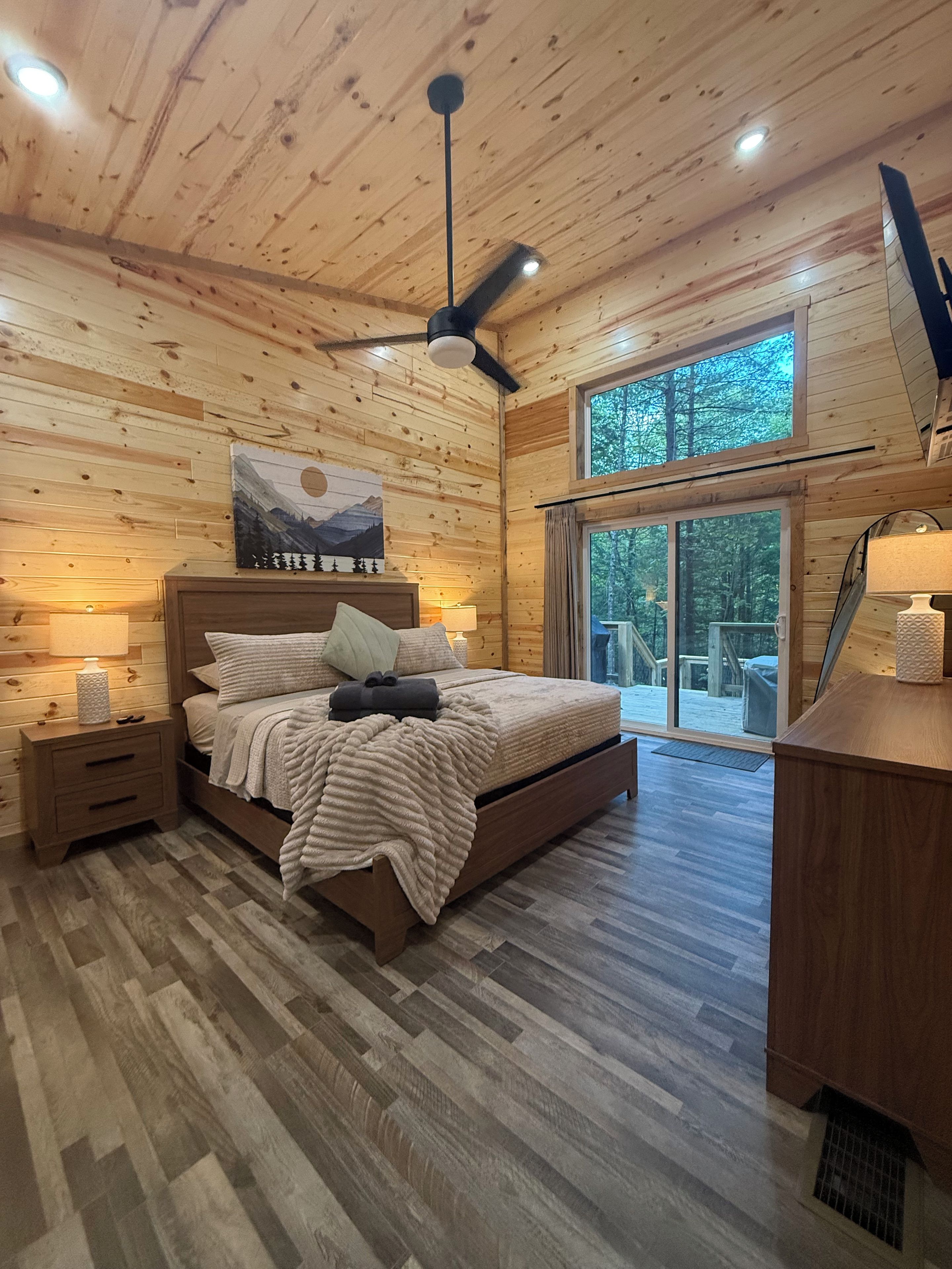 A wooden cabin bedroom with a bed, nightstands, a ceiling fan, and a large window.