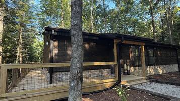 A wooden cabin with a deck, surrounded by trees and a clear sky.