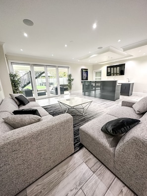 A modern living room with a large sectional sofa, a glass coffee table, and a kitchen area with a flat-screen TV. - Luxury Large Private Modern Home with Pool (West Vancouver)