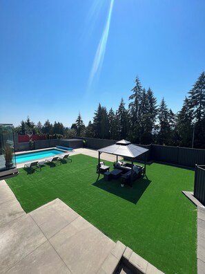 A backyard with a swimming pool, artificial grass, a gazebo, and a patio area.