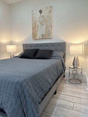 A bedroom with a large bed, two bedside lamps, a grey upholstered headboard, and a painting on the wall. - Luxury Large Private Modern Home with Pool (West Vancouver)