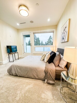 A bedroom with a large bed, a TV, a window with a view of trees, and a painting on the wall.