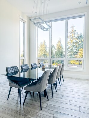 A modern dining area with a glass table, grey upholstered chairs, and large windows offering a view of trees. - Luxury Large Private Modern Home with Pool (West Vancouver)