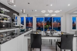 A modern kitchen with a large island, black chairs, and a view of trees through large windows. - Luxury Large Private Modern Home with Pool (West Vancouver)
