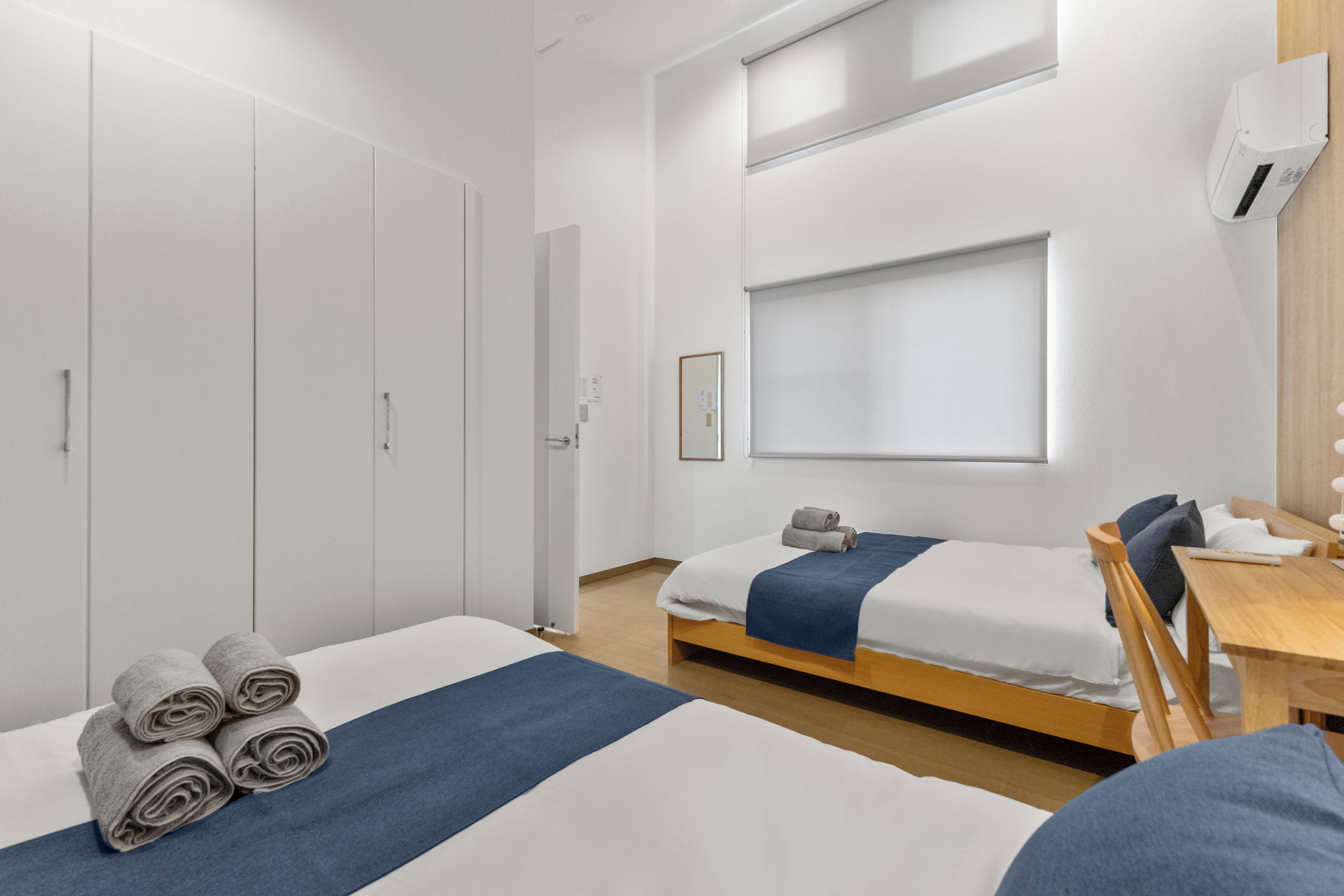 Habitación de hotel con dos camas, un escritorio, una silla, un armario y una ventana con persianas.