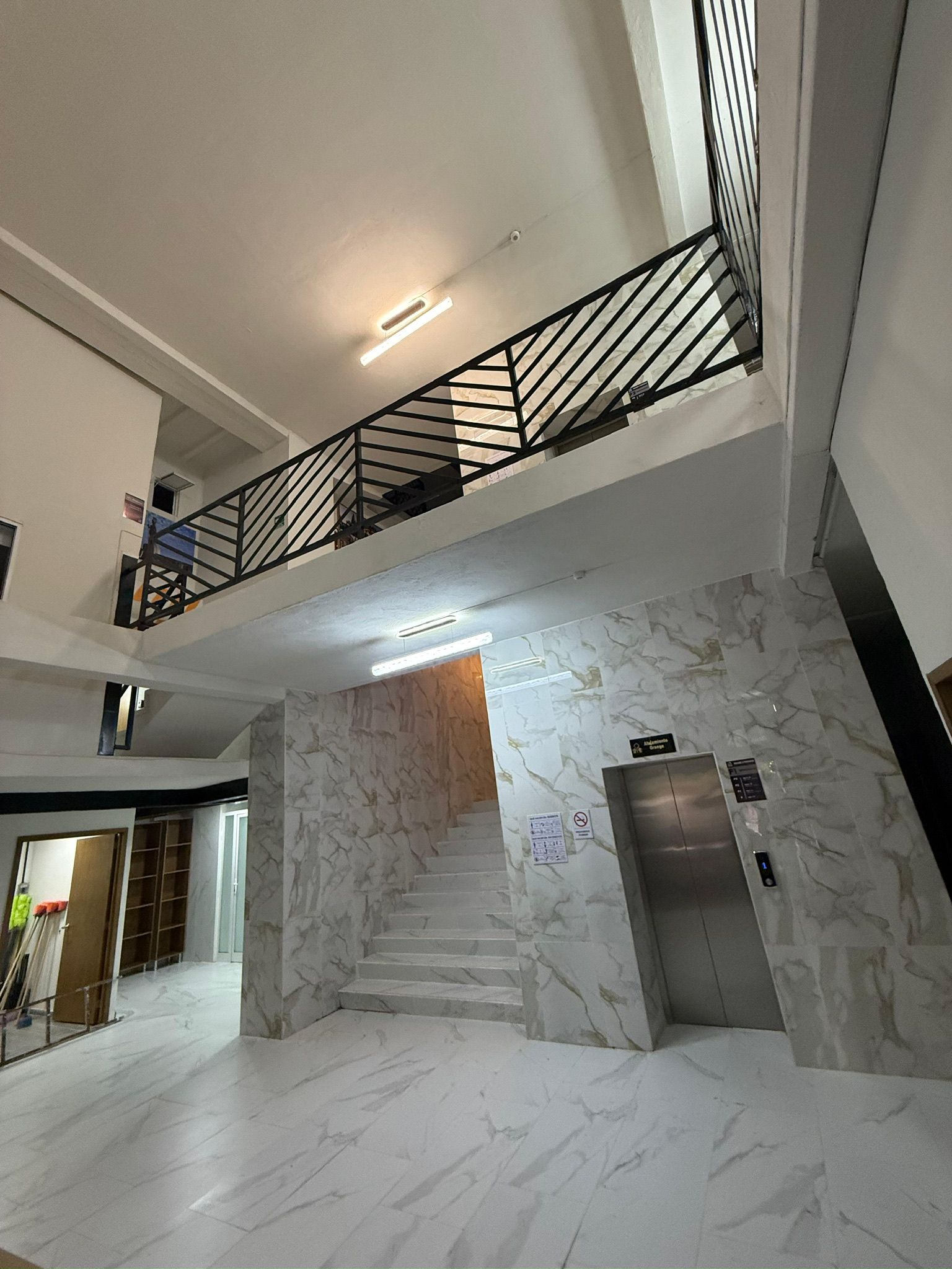 Un interior espacioso con una escalera de mármol, un balcón en el segundo piso con barandilla metálica y un ascensor.