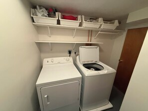 A laundry room with a front-loading washer and dryer, a shelf above with storage bins, and a door to the right. - 3-bedroom apartment in peaceful Tremonton (Tremonton)