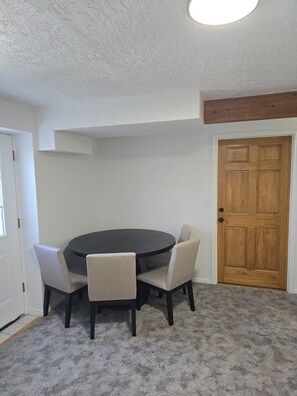 Um quarto com uma mesa redonda e quatro cadeiras, uma porta de madeira e teto branco.