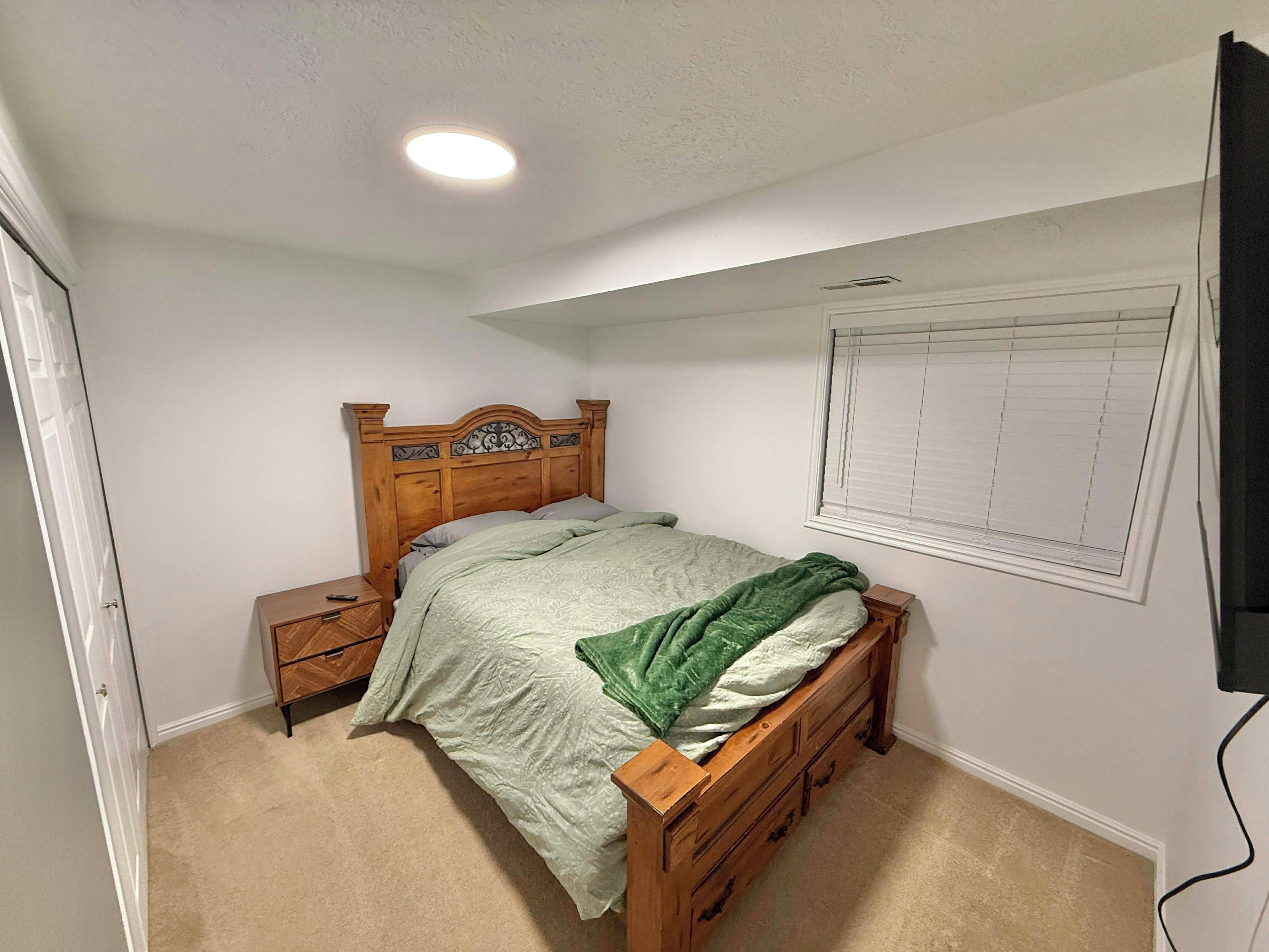 Une chambre avec un lit simple, une table de chevet, une commode, une fenêtre avec des stores et un luminaire au plafond.