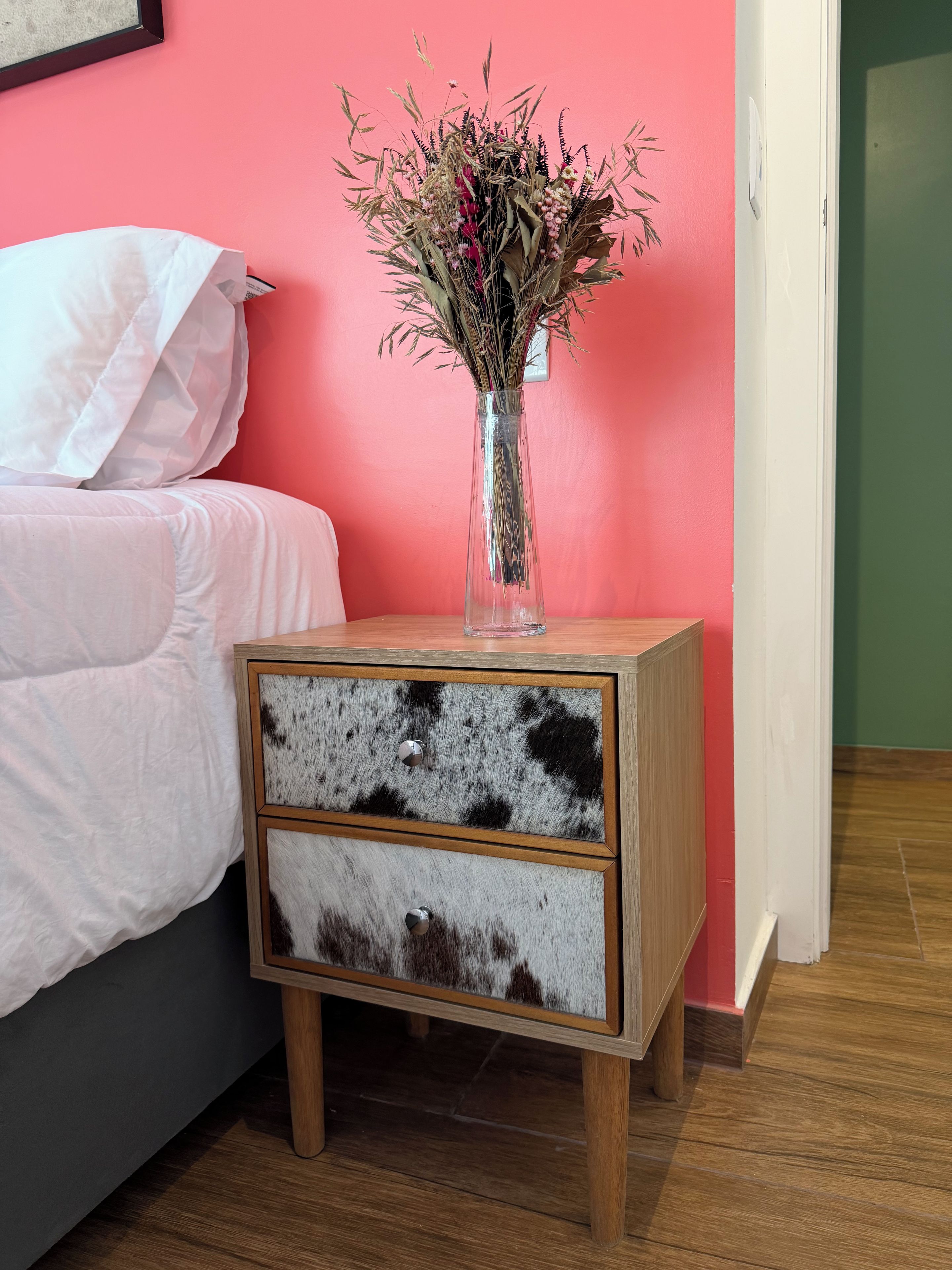 Uma mesa de cabeceira com um vaso de flores secas, uma cama com roupa de cama branca e uma parede rosa.
