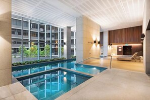 A modern indoor swimming pool with clear glass walls, a lounge area with seating, and a view of a building with balconies. - Melbourne Skyline Views Modern 1BR with Pool & Gym (Melbourne)
