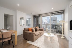A modern living room with a sofa, a dining table set for two, and a large window offering a city view. - Melbourne Skyline Views Modern 1BR with Pool & Gym (Melbourne)