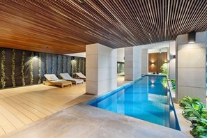 A modern indoor swimming pool with lounge chairs, a wooden ceiling, and a stone floor. - Melbourne Skyline Views Modern 1BR with Pool & Gym (Melbourne)