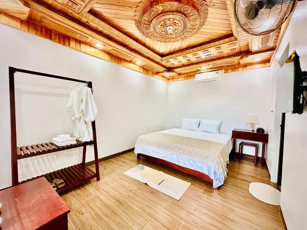 Habitación de hotel con techo de madera, cama con cubrecama blanco, mesita de noche con lámpara y banco de madera con toallas.