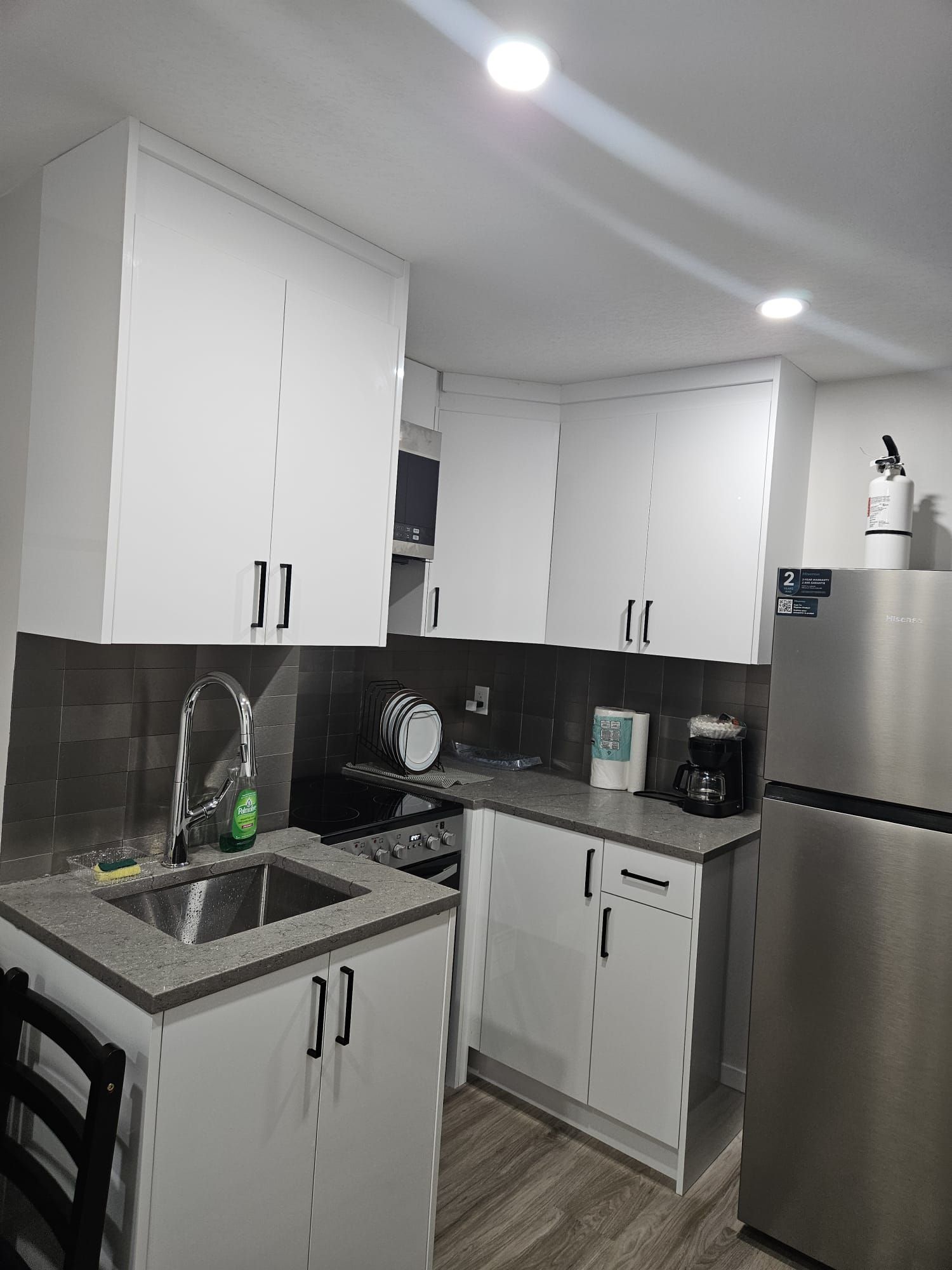 A modern kitchen with white cabinets, a stainless steel refrigerator, a coffee maker, and a sink with a dish rack.