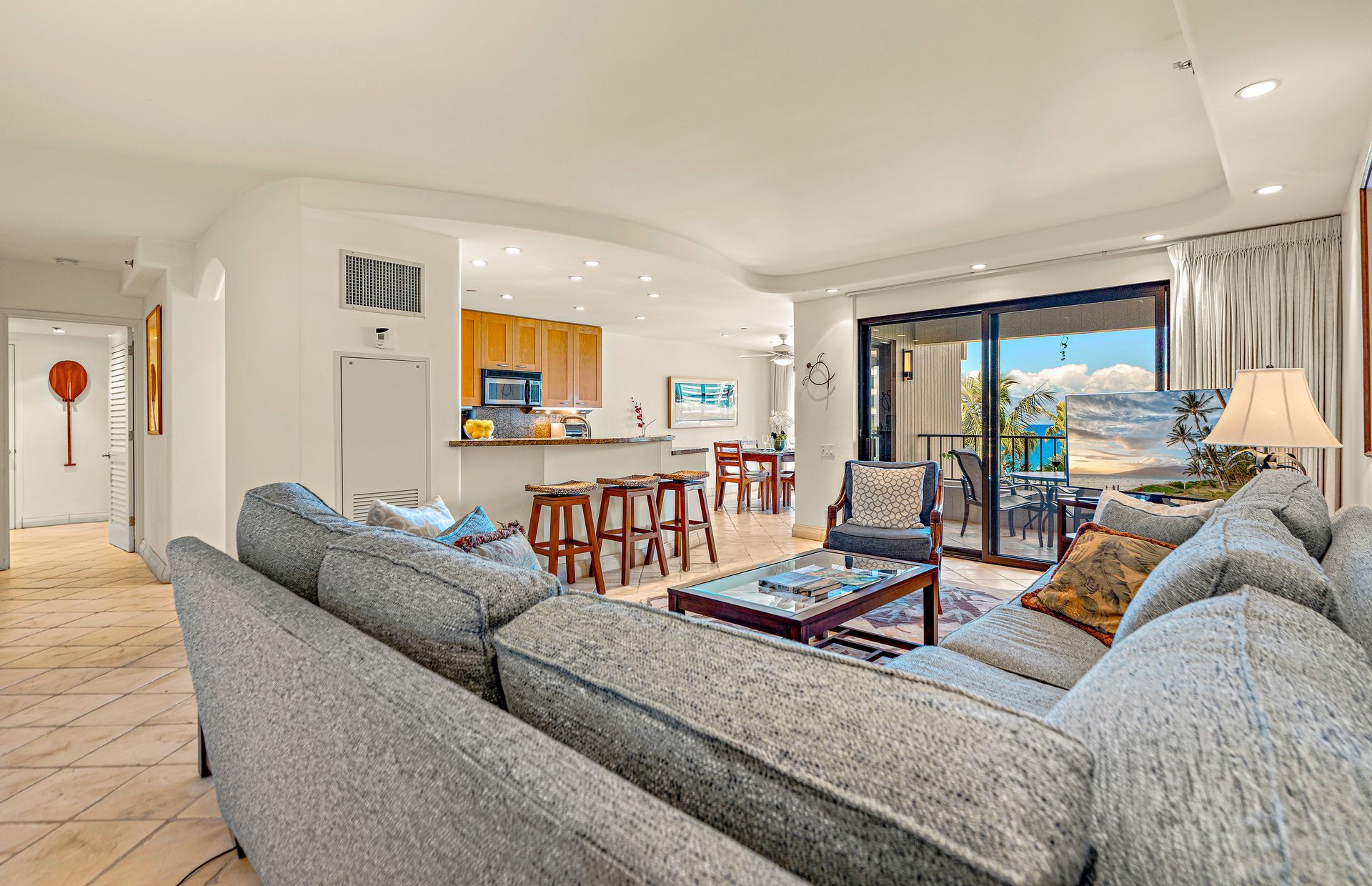 A modern living room with a grey sofa set, a dining area with wooden chairs, a kitchen with wooden cabinets, and a balcony with a view of palm trees.